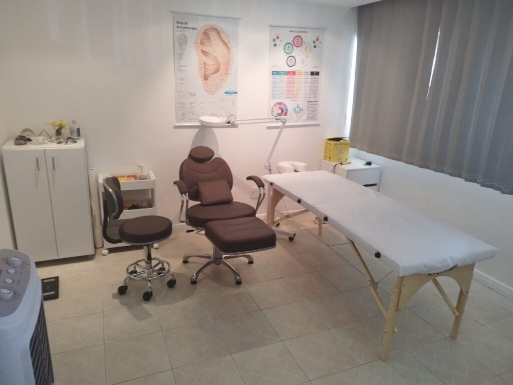 Sala de atendimento do Espaço Terapêutico Maura Chiattone, com maca, cadeira de auriculoterapia e mapas de pontos da orelha Sala de atendimento do Espaço Terapêutico Maura Chiattone, com maca, cadeira de auriculoterapia e mapas de pontos da orelha